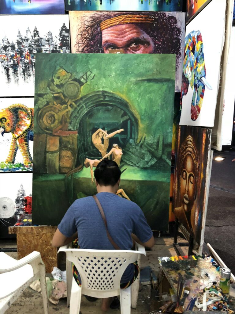 An artist sits indoors, painting a surreal artwork amidst other colorful canvases at an exhibition.